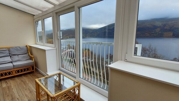 A balcony with a sofa and a glass table, overlooking a lake and hills. - Red Deer Lodge 06 Loch Rannoch Highland Club (Pitlochry)