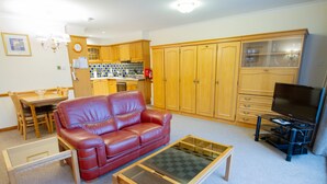 Ein Wohnzimmer mit einem roten Ledersofa, einem Glastisch, Holzschränken, einem Fernseher und einer Küche im Hintergrund.