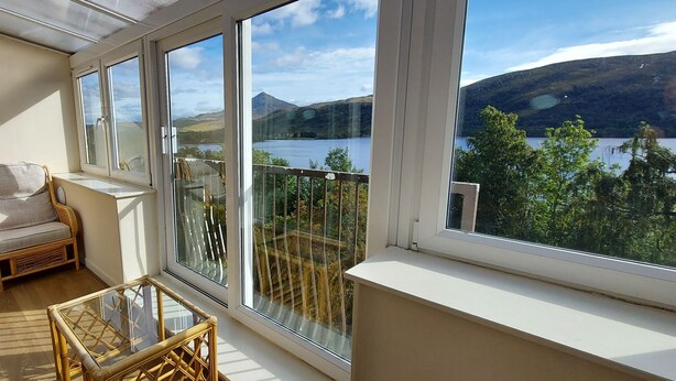 Ein Balkon mit Korbmöbeln, einem Glastisch und Blick auf einen See und Berge.