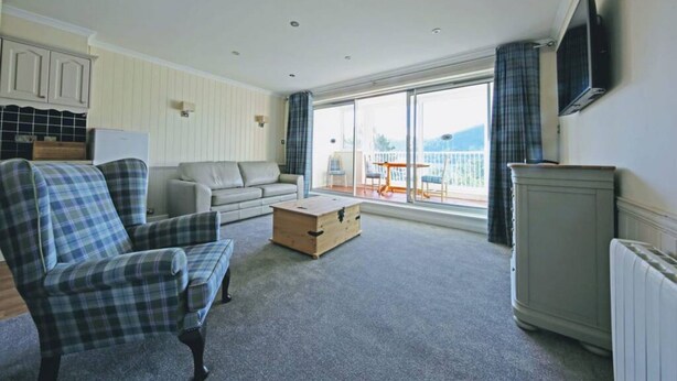 A living room with a sofa, armchair, and a wooden chest. There is a television mounted on the wall, a window with curtains, and a view of the outdoors. - Apartment 09 Loch Rannoch Highland Club (Pitlochry)