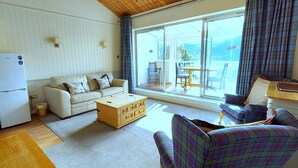 A compact living space with a kitchenette, a sofa, a plaid armchair, and a view through large windows. - Apartment 10 Loch Rannoch Highland Club (Pitlochry)
