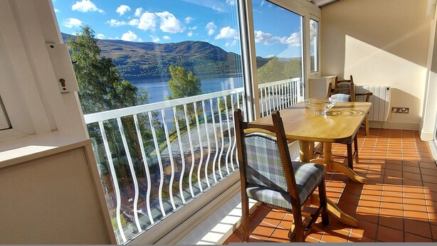 A balcony with a view of a lake and mountains, a wooden table with chairs, and a window with a white frame. - Apartment 10 Loch Rannoch Highland Club (Pitlochry)