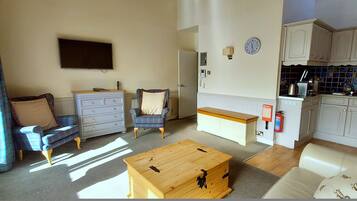 A compact living space with a sofa, a wooden coffee table, a chair, a dresser, a television, a kitchen area with cabinets and a tiled backsplash, and a window with curtains.