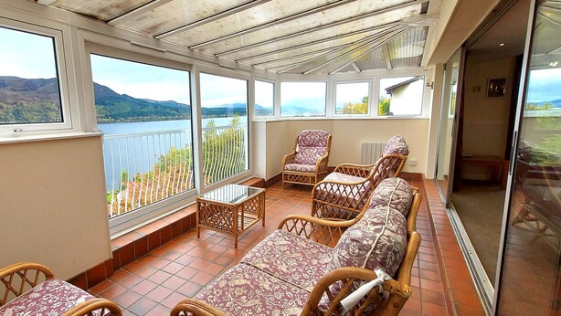 A sunlit balcony with wicker furniture, large windows offering a view of a lake and hills. - Highland Lodge 36 Loch Rannoch Highland Club (Pitlochry)