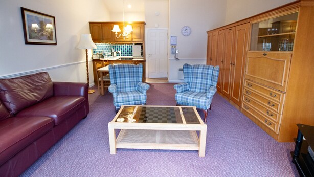A living room with a red sofa, two plaid armchairs, a coffee table, a wooden cabinet, and a kitchen area in the background. - Studio 05 Loch Rannoch Highland Club (Pitlochry)