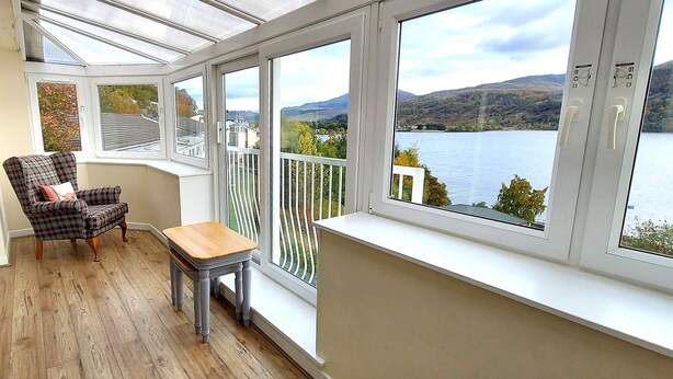 A conservatory with a wooden floor, a plaid chair, a small table, and a view of a lake and hills. - Pine Marten Lodge 08 Loch Rannoch Highland Club (Pitlochry)