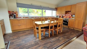 A kitchen with wooden cabinets, a dining table with chairs, a window with a view of greenery, and a microwave. - Highland Lodge 02 Loch Rannoch Highland Club (Pitlochry)