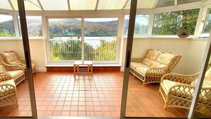 A room with wicker furniture, a tiled floor, and large windows offering a view of hills and a lake. - Highland Lodge 02 Loch Rannoch Highland Club (Pitlochry)