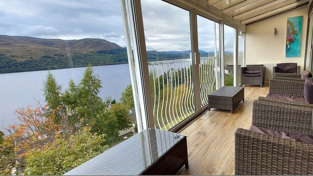 A balcony with wicker furniture, a glass railing, and a view of a lake and hills. - Highland Lodge 35 Loch Rannoch Highland Club (Pitlochry)