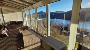 A covered outdoor area with wicker furniture, large windows, and a view of a lake and mountains. - Highland Lodge 35 Loch Rannoch Highland Club (Pitlochry)