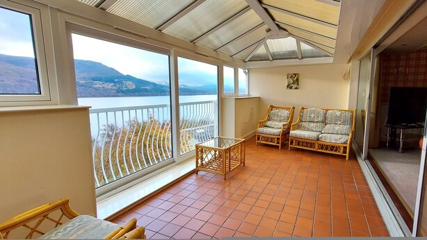 A balcony with wicker furniture, a TV, and a view of a lake and mountains. - Highland Lodge 46 Loch Rannoch Highland Club (Pitlochry)