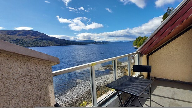A balcony with a metal table and chair, overlooking a lake with mountains in the distance. - Lochside Lodge 03 Loch Rannoch Highland Club (Pitlochry)
