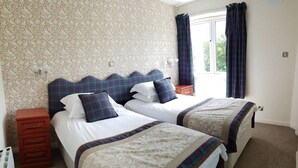 A double room with two beds, each with a plaid headboard and matching pillows. There is a wooden nightstand with drawers and a small table beside the beds. The room has a patterned wallpaper and a window with curtains. - Highland Lodge 38 Loch Rannoch Highland Club (Pitlochry)