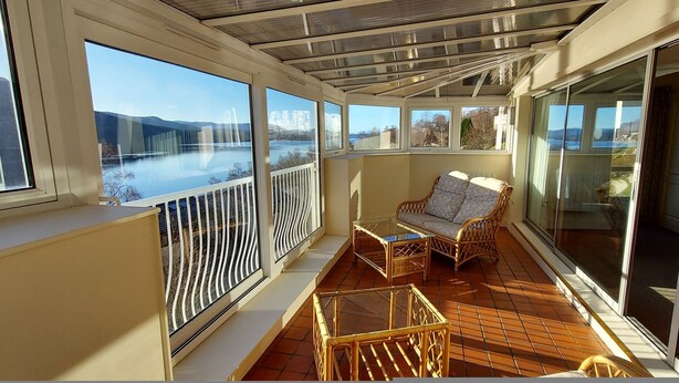 A sunlit balcony with a view of a lake, wicker furniture, and a glass door leading to the interior. - Highland Lodge 38 Loch Rannoch Highland Club (Pitlochry)