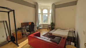 Ein Schlafzimmer mit einem Bett, einem Schreibtisch mit Computer, einem roten Sofa und einem Fenster mit Vorhängen.