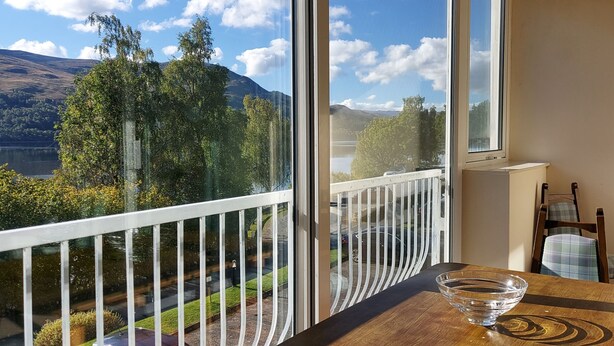 Een balkon met vrij uitzicht op een meer en bergen, een houten tafel met een glazen schaal, en een stoel met een geruit kussen.