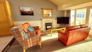 A living room with a fireplace, a television, and a balcony with yellow chairs. - Highland Lodge 04 Loch Rannoch Highland Club (Pitlochry)