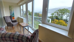A balcony with a view of a lake and mountains, a plaid sofa, a chair, and a small table.