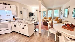 A modern kitchen with white cabinets, a granite countertop, and a built-in oven. There is a dining area with a white table and chairs, and a living room with a sofa and a coffee table. - Windmill Cottage at Abbey Springs Near Lake & Shops! (Fontana)