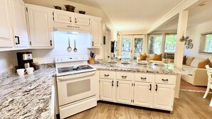 A kitchen with white cabinets, a stove, and a coffee maker. There is a dining area with a table, chairs, and a view of the outdoors. - Windmill Cottage at Abbey Springs Near Lake & Shops! (Fontana)