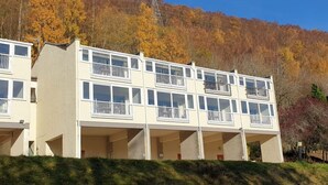 Un edificio de varios pisos con balcones, frente a un paisaje de árboles de colores otoñales.