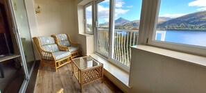 A balcony with wicker furniture and a glass table, overlooking a lake and mountains.