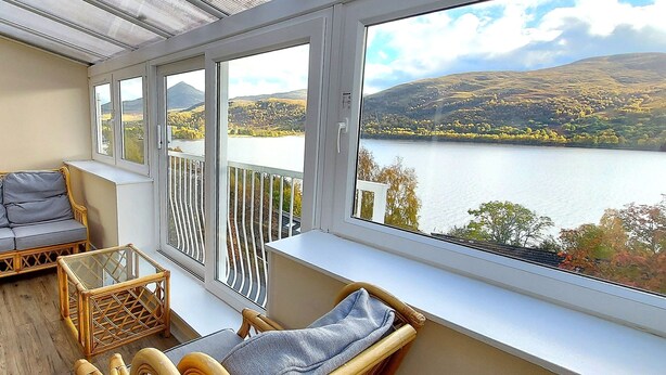 Ein Balkon mit einer Couch, einem kleinen Tisch und Korbstühlen mit Blick auf einen See und Hügel.