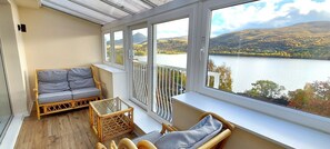 Un balcon avec un canapé, une petite table et des chaises en osier, donnant sur un lac et des collines.