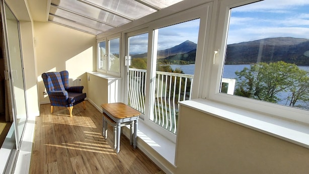 A balcony with a view of a lake and mountains, a blue plaid chair, and a small wooden table. - Pine Marten Lodge 02 Loch Rannoch Highland Club (Pitlochry)