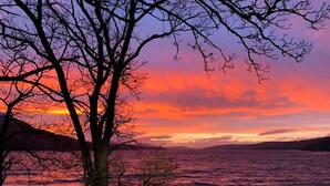 Ein Baum im Vordergrund, im Hintergrund ein Sonnenuntergang über einem Gewässer.