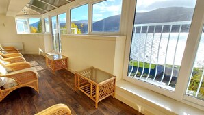 Ein Zimmer mit Korbmöbeln, einem Glastisch und großen Fenstern mit Blick auf ein Gewässer und Hügel.
