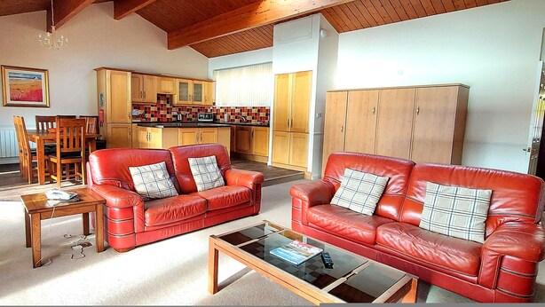 Ein Wohnzimmer mit roten Ledersofas, einem Holztisch und einem Couchtisch mit Glasplatte. Im Hintergrund befindet sich eine Essecke mit Holzkästen und einem Tisch, sowie eine Küche.