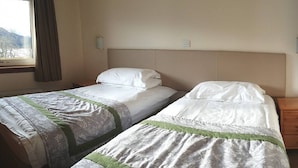 Ein Hotelzimmer mit zwei Einzelbetten, einem Fenster mit Vorhängen, einem Nachttisch aus Holz und einer Wandlampe.