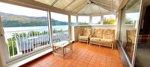 A sunroom with large windows overlooking a lake and hills, furnished with wicker chairs and a glass table. - Highland Lodge 42 Loch Rannoch Highland Club (Pitlochry)