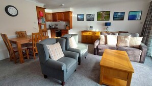 A living room with a grey armchair, a brown sofa, a wooden coffee table, and a dining area with wooden chairs and a table. - Red Deer Lodge 04 Loch Rannoch Highland Club (Pitlochry)