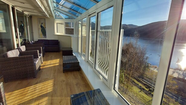 A sunlit room with wicker furniture, large windows, and a view of a lake and hills. - Highland Lodge 06 Loch Rannoch Highland Club (Pitlochry)