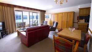 A living room with a red sofa, a wooden dining table with a bowl, and a view of a balcony through large windows. - Otter Lodge 08 Loch Rannoch Highland Club (Pitlochry)