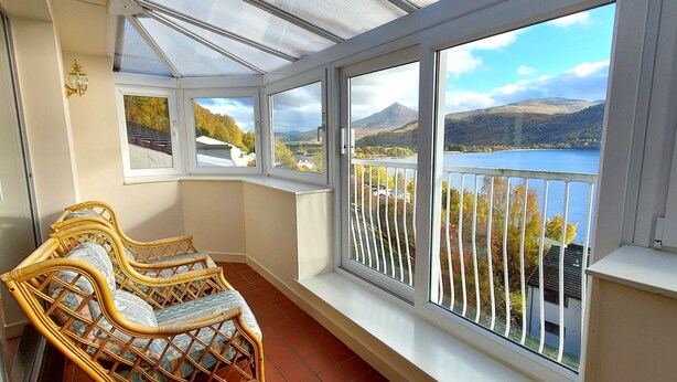 A room with a bay window overlooking a lake and mountains, featuring wicker chairs and a glass ceiling. - Otter Lodge 08 Loch Rannoch Highland Club (Pitlochry)
