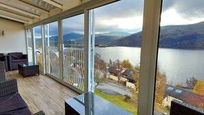 Ein Balkon mit Korbmöbeln, große Fenster mit Blick auf einen See und Berge.