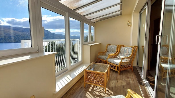 A balcony with wicker furniture, a glass table, and a view of a lake and mountains. - Otter Lodge 06 Loch Rannoch Highland Club (Pitlochry)