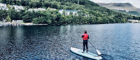 En person står på paddleboard på en rolig sø, med en skovklædt bakke i baggrunden.