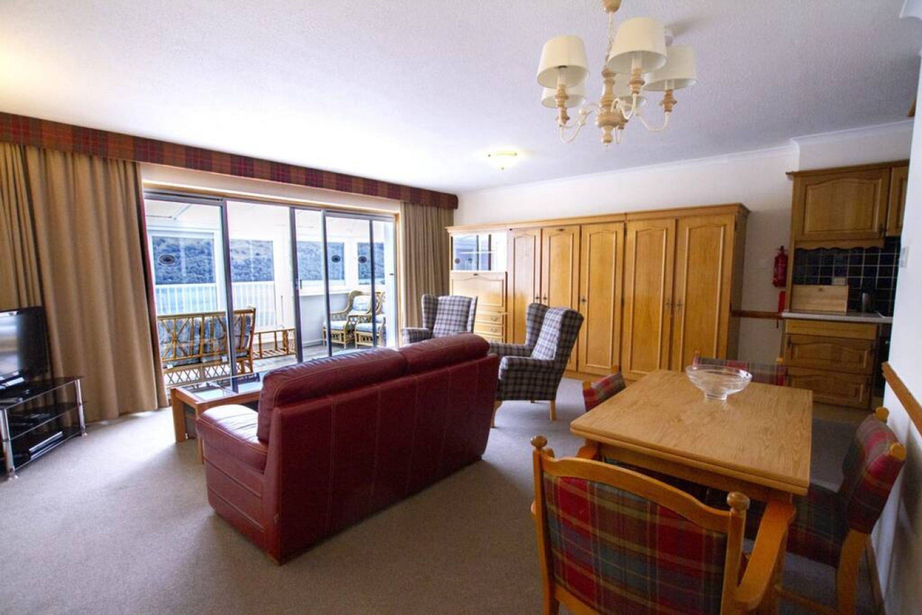 A living room with a red sofa, a wooden dining table with a bowl, and a view of a balcony through large windows.