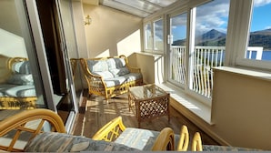 A sunlit balcony with wicker furniture, a glass table, and a view of a lake and mountains. - Otter Lodge 06 Loch Rannoch Highland Club (Pitlochry)