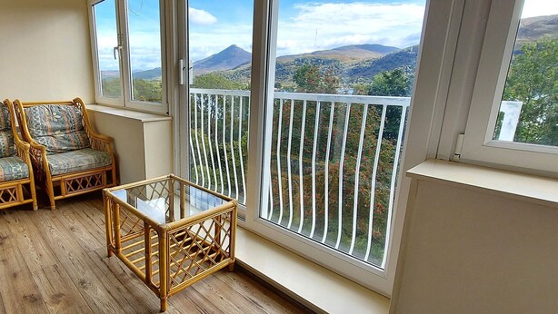 A balcony with wicker chairs and a glass table, offering a view of distant hills and trees. - Red Squirrel Lodge 03 Loch Rannoch Highland Club (Pitlochry)