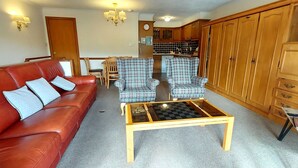 A living room with a red leather sofa, plaid armchairs, a wooden coffee table, and a kitchen in the background. - Red Squirrel Lodge 03 Loch Rannoch Highland Club (Pitlochry)