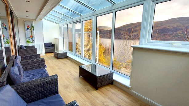 A spacious room with a large window overlooking a lake and hills. There are wicker sofas and a glass coffee table. - Highland Lodge 07 Loch Rannoch Highland Club (Pitlochry)