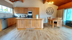 A modern kitchen with wooden cabinets, a central island, and a dining area with wooden chairs. - Highland Lodge 07 Loch Rannoch Highland Club (Pitlochry)