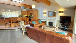A spacious living room with a brown leather sofa, a coffee table, and a fireplace. There is a dining area with wooden chairs and a kitchen in the background.