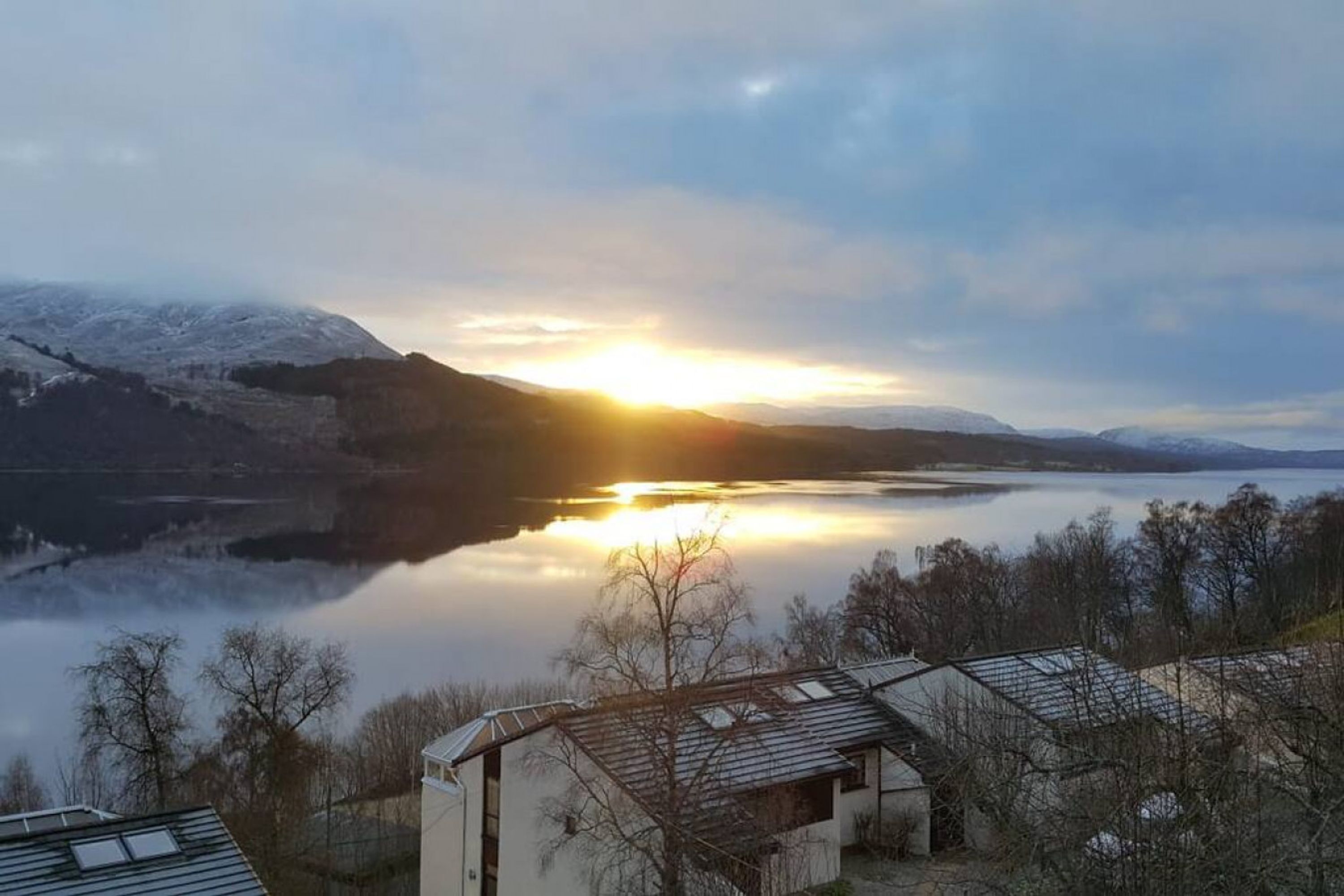 A lakeside view with a building in the foreground, a sunset in the background, and snow-covered hills.