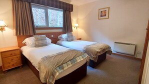 A bedroom with two beds, a nightstand, a window with curtains, wall-mounted lamps, and a radiator. - Otter Lodge 04 Loch Rannoch Highland Club (Pitlochry)